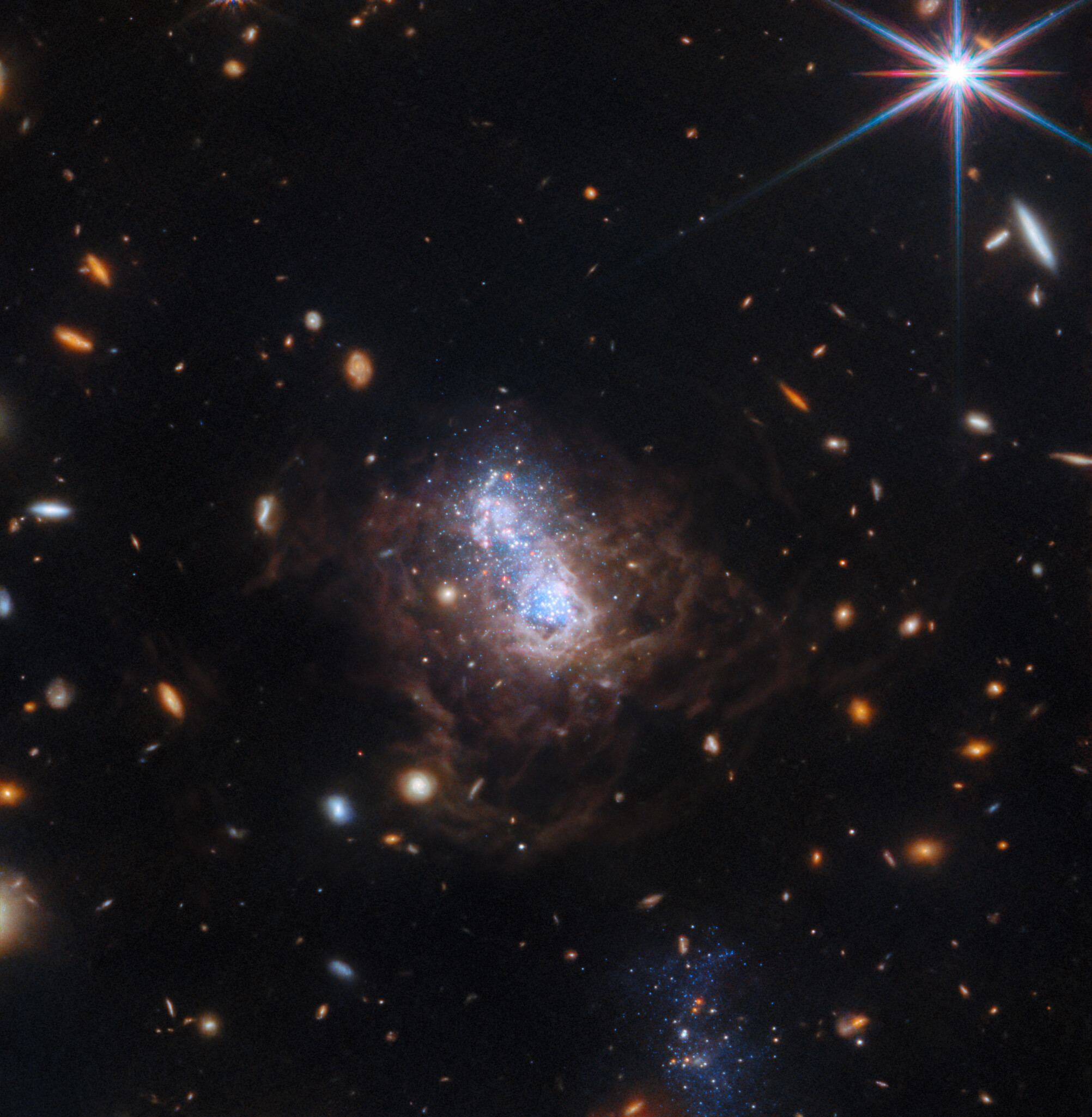 Na ilustracji: W polu widzenia widać wiele małych galaktyk. Najczęściej są to owalne galaktyki spiralne w kolorze białym i czerwonym. Centrum zdjęcia wypełnia nieregularna galaktyka karłowata I Zwicky 18 (I Zw 18) z jasnym obszarem wypełnionym gwiazdami w kolorze białym i niebieskim. Jądro galaktyki rozdziela się na dwa płaty. Obszar centralny I Zw 18 jest otoczony przez włókna pyłowe w kolorze brązowym. W dolnej części zdjęcia jest widoczna galaktyka towarzysząca (wygląda jak zbiór niebieskich gwiazd), która najprawdopodobniej oddziałuje z I Zw 18 i mogła w niej zapoczątkować niedawne procesy gwiazdotwórcze.  ESA/Webb, NASA, CSA, A. Hirschauer, M. Meixner et al.