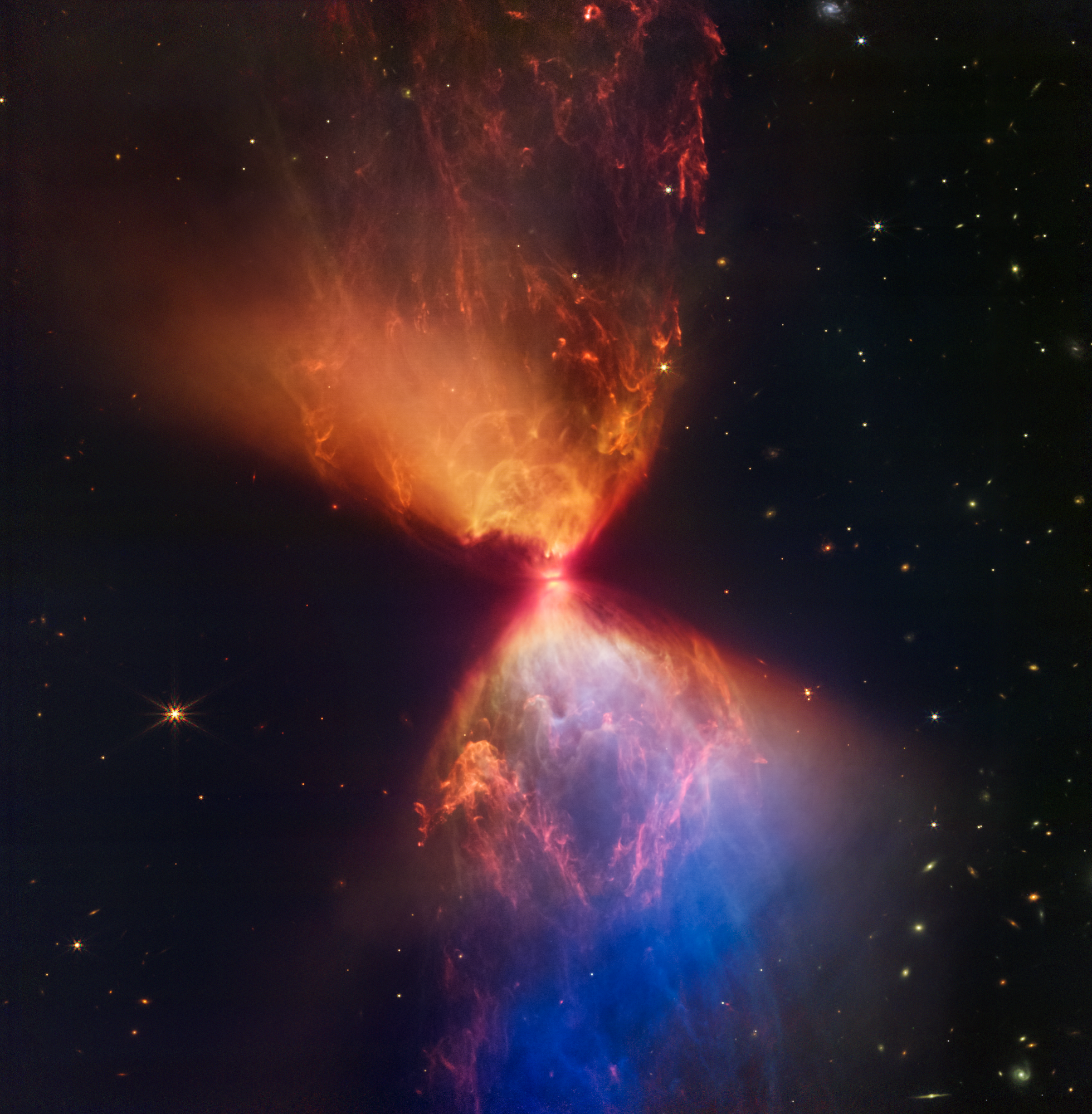 Na zdjęciu w bliskiej podczerwieni wykonanym przez Kosmiczny Teleskop Jamesa Webba (kamera NIRCam) widać protogwiazdę w ciemnej mgławicy L1527. Protogwiazda jest zanurzona w chmurze materii, którą akreuje i przez to staje się coraz cięższa. Wyrzuty materii z protogwiazdy wyczyściły przestrzeń powyżej i poniżej tego ciemnego obłoku, którego granice mają barwę pomarańczową i niebieską. W górnym, centralnym obszarze widać struktury podobne do bąbli, które powstały w wyniku sporadycznych wyrzutów materii. Teleskop Webba zarejestrował również włókna składające się z molekularnego wodoru, które zostały zjonizowane przez wyrzuconą materię w przeszłości. Brzegi obszarów z wydmuchaną materią u góry po lewej i na dole po prawej są proste, natomiast granice obszarów u góry po prawej i na dole po lewej są zakrzywione. Obszar na dole po prawej jest niebieski, ponieważ jest mniej pyłu pomiędzy tym obszarem i teleskopem Webba w porównaniu do obszaru pomarańczowego powyżej protogwiazdy. Źródło: NASA, ESA, CSA, STScI, Joseph DePasquale (STScI), Alyssa Pagan (STScI), Anton M. Koekemoer (STScI)