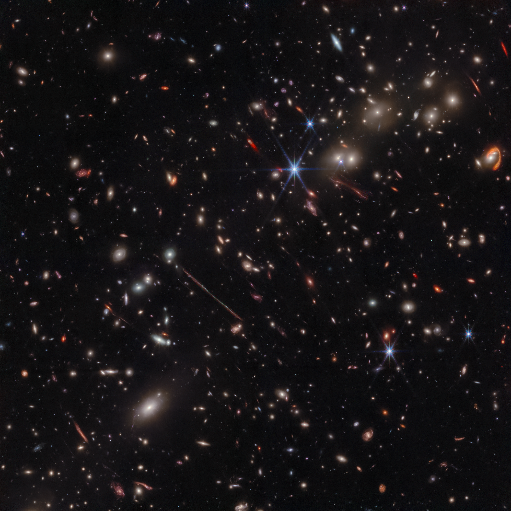 Na ilustracji: Zdjęcie w podczerwieni gromady galaktyk El Gordo („Grubas”) zawierającej setki galaktyk, które zostało zrobione przez Teleskop Webba. El Gordo działa jak soczewka grawitacyjna zniekształcająca i wzmacniająca światło odległych galaktyk tła. Najciekawsze z nich to zniekształcona do kreski galaktyka tła La Flaca („Chuda”) widoczna nieco na lewo od środka zdjęcia oraz El Anzuelo („Haczyk”) widoczna w pobliżu górnego prawego narożnika zdjęcia.  Źródło: NASA, ESA, CSA