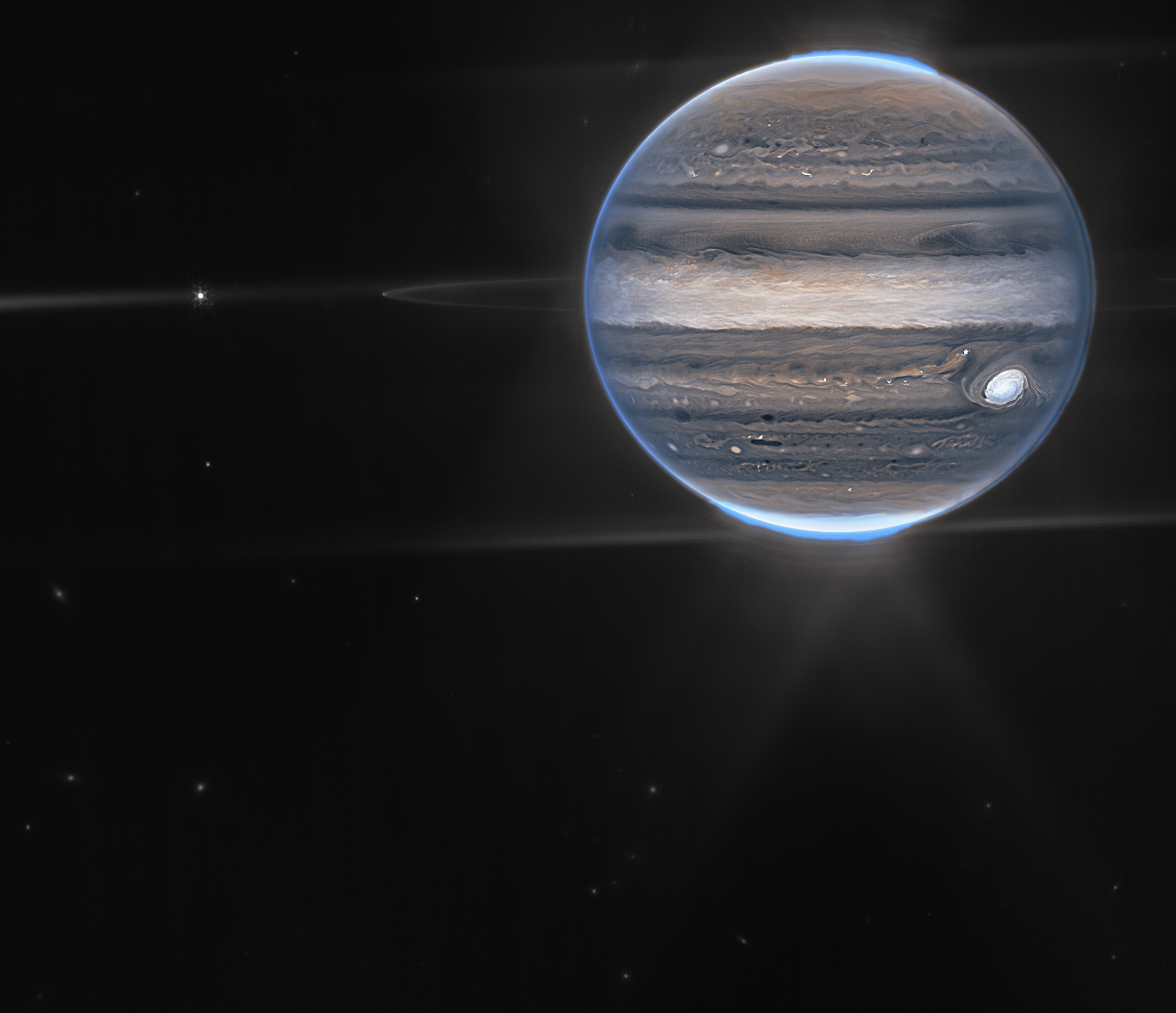 Na ilustracji: Zdjęcie Jowisza zrobione Teleskopem Webba z kamerą NIRCam  – w dwóch barwach / filtrach: F212N (pomarańczowy) i F335M (cyjan/niebieskozielony)  – oraz w dwóch wersjach: bez etykiet (u góry), z etykietami (na dole). Na zdjęciu widać szczegóły atmosfery Jowisza (np. gigantyczne burze, potężne wiatry, zorze polarne), jego pierścienie (około milion razy słabsze niż planeta), niektóre małe księżyce (Amaltea, Adrastea), a nawet galaktyki w tle (rozmyte plamki). Słynna burza – Wielka Czerwona Plama, trwająca w atmosferze Jowisza już od co najmniej 400 lat, wydaje się biała na zdjęciu – podobnie jak inne chmury. Jest to spowodowane odbijaniem dużej ilości światła słonecznego. W przeciwieństwie do tego, ciemne pasma na północ od obszaru równikowego mają małe pokrycie chmurami. Na tym zdjęciu jeżeli coś jest jasne, to znaczy, że jest wysoko w atmosferze Jowisza. Dlatego Wielka Czerwona Plama posiada takie zamglenia na dużej wysokości – podobnie jak  obszar równikowy. Liczne jasne, białe plamy i pasemka są najprawdopodobniej bardzo wysokimi wierzchołkami chmur z burz konwekcyjnych. Źródło: NASA, ESA, CSA, Jupiter ERS Team; opracowanie zdjęcia Ricardo Hueso (UPV/EHU) i Judy Schmidt (projekt nauki obywatelskiej)