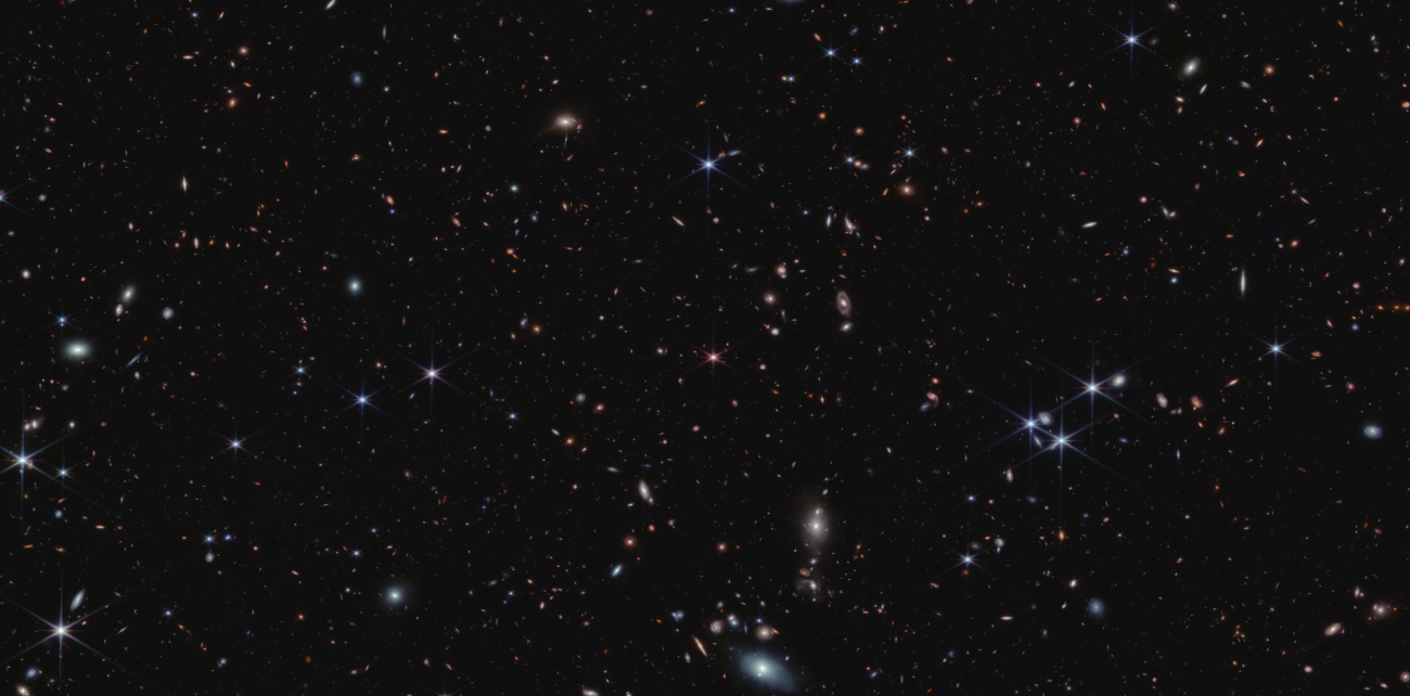 W centrum ilustracji znajduje się kwazar J0100+2802 (z=6,3)–różowy obiekt z ośmioma „spajkami”, będącymi znakiem rozpoznawczym Teleskopu Webba (gwiazdy tła mają te spajki dłuższe i w kolorze niebieskim). Na tym zdjęciu w podczerwieni na pograniczu konstelacji Ryb i Andromedy widać również tysiące różnokolorowych galaktyk. Niektóre z tych najmniejszych są w odcieniach pomarańczowych lub różowych. Najbardziej odległe galaktyki wyglądają jak pojedyncze kropki świetlne. Źródło: NASA, ESA, CSA, Simon Lilly i in.