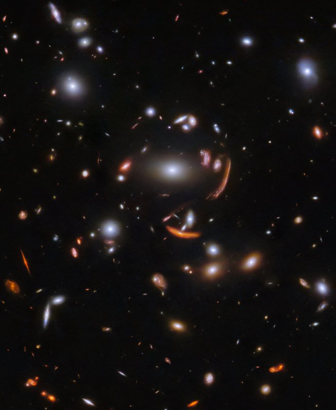 Na ilustracji: Gromada galaktyk SDSS J1226+2152 sfotografowana w podczerwieni przez Teleskop Webba. Większość galaktyk na zdjęciu ma kształt owalny i gładki. Kilka galaktyk posiada ramiona spiralne nachylone pod różnymi kątami. Największa galaktyka znajduje się w samym centrum zdjęcia i jest otoczona przez kilka obrazów galaktyk tła rozciągniętych i wygiętych w długie łuki przez soczewkowanie grawitacyjne. Tło jest czarne i zawiera wiele drobnych galaktyk, ale żadnej gwiazdy! Źródło: ESA/Webb, NASA & CSA, J