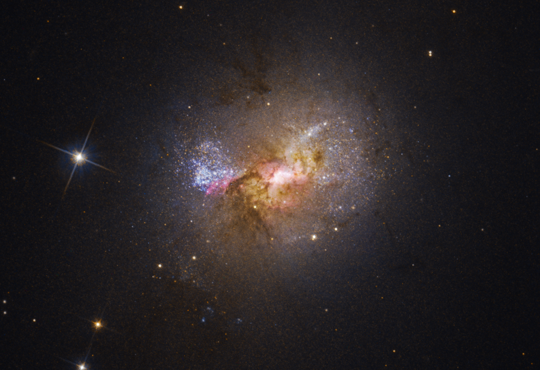 Zdjęcie robione przez Kosmiczny Teleskop Hubble’a w zakresie optycznym galaktyki karłowatej Henize 2-10 mieniącej się młodymi gwiazdami. Jasny obszar w centrum otoczony przez różowe obłoki i pasy ciemnego pyłu wskazują na położenie masywnej czarnej dziury i aktywnych obszarów tworzenia się gwiazd. Źródło: NASA, ESA, Zachary Schutte (XGI), Amy Reines (XGI), Alyssa Pagan (STScI)