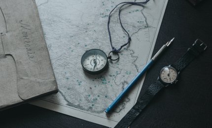 Niebiesko-białe pióro leżące obok srebrnego okrągłego zegarka, starej mapy i kompasu