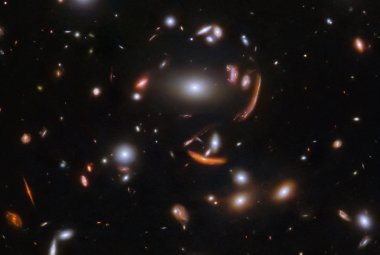 Na ilustracji: Gromada galaktyk SDSS J1226+2152 sfotografowana w podczerwieni przez Teleskop Webba. Większość galaktyk na zdjęciu ma kształt owalny i gładki. Kilka galaktyk posiada ramiona spiralne nachylone pod różnymi kątami. Największa galaktyka znajduje się w samym centrum zdjęcia i jest otoczona przez kilka obrazów galaktyk tła rozciągniętych i wygiętych w długie łuki przez soczewkowanie grawitacyjne. Tło jest czarne i zawiera wiele drobnych galaktyk, ale żadnej gwiazdy! Źródło: ESA/Webb, NASA & CSA, J